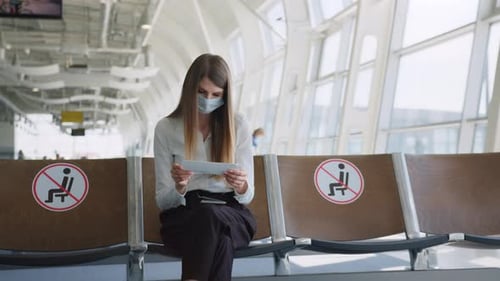 Confident Business Woman in a Mask Sitting at the Airport Holding a Passport and