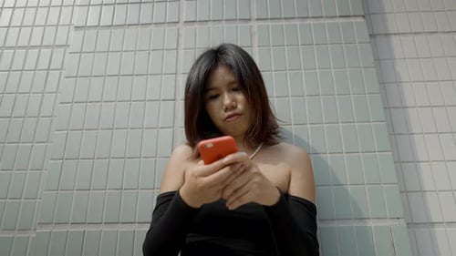 Asian Woman in Black Sexy Dress with Smartphone Standing Against Building Background