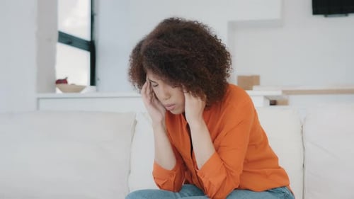 Unhappy African American Woman Sit on Couch at Home Kitchen Living Room Rubs Temple Feeling Pain