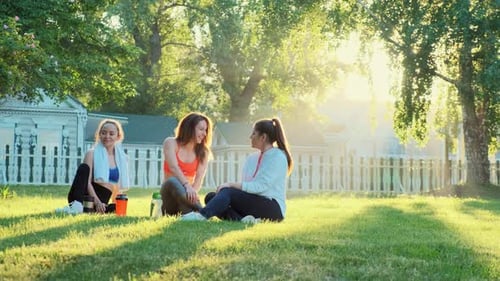 Three Girlfriends Are Sitting in a Clearing of Green Grass After Jogging Laughing and Talking.