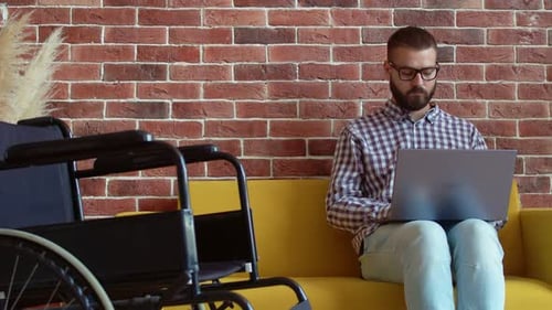 Man Using Laptop on Sofa Beside Wheelchair
