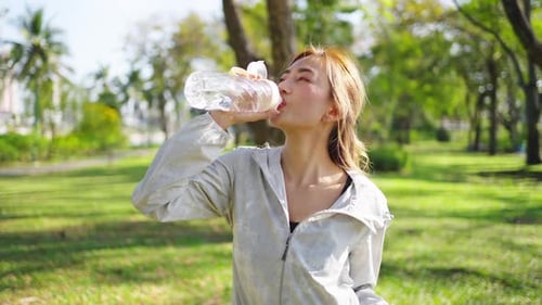 4K Asian woman drinking water from a bottle while jogging at public park in the morning.