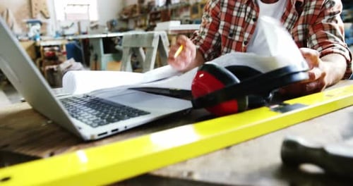 Man Reviews Blueprints in Workshop With Laptop