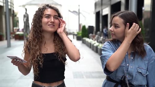 Girlfriends Are Walking Along a City Street and Chatting. One Curly-haired Woman Holds a Phone and