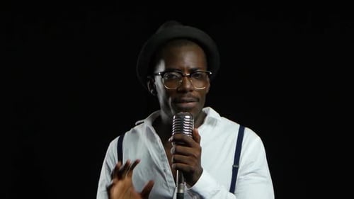 Man Singer canta en un micrófono, baila y chasquea los dedos. Fondo negro. De cerca