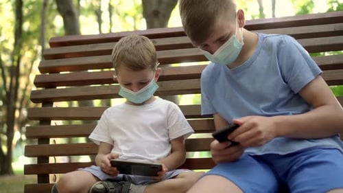 Boys in Medical Masks Play with Smartphones in the Park