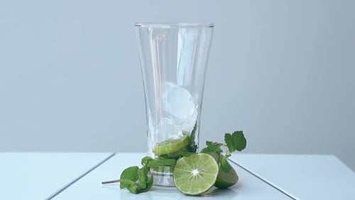 Glass with Lime and Ice Cubes Stands on White Table at Wall