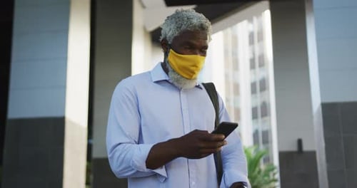 Hombre mayor afroamericano con máscara facial usando un teléfono inteligente y mirando a su alrededor en un parque Corporativa