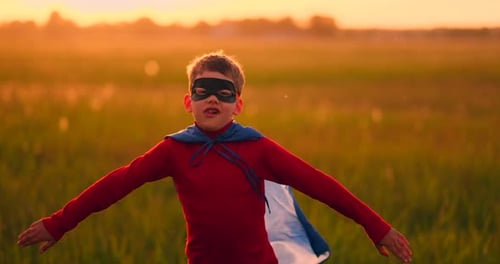 A Boy in a Suit and a Superhero Mask Running Across the Field at Sunset on the Grass