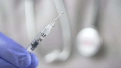 Doctor with a Phonendoscope and Blue Gloves Offers a Syringe with a Vaccine