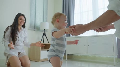 Happy family, mother support young toddler son taking first step walk on floor to father in house.