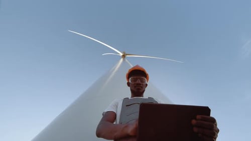Afrikanischer Ingenieur im Overall steht zwischen Windmühlenfarm mit modernem Tablet
