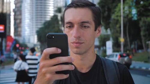 Portrait of Tourist Man with Backpack Using Smartphone Standing Outdoors Alone Smiling at Urban City
