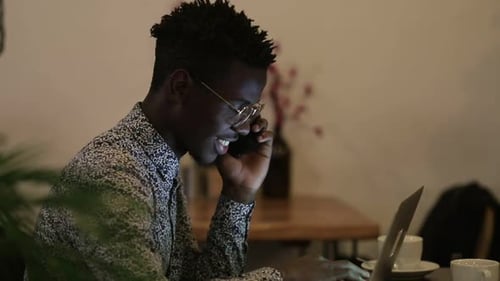 Smiling Young Man Talking By Smartphone and Using Laptop in Cafe