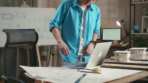 Asian Man Engineer Walk Into The Office Before Typing On A Laptop And Looking At Blueprint