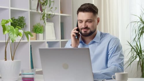 A young friendly man with a beard sitting in the office talking on a mobile phone