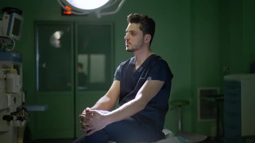 Side View Portrait of Tired Young Male Surgeon Sitting in Operating Room on Medical Couch Thinking