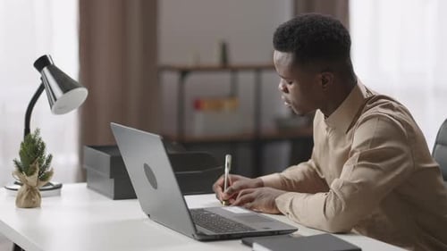 Young Afroamerican Businessman is Working at Home Sitting at Table with Laptop and Writing Notes in