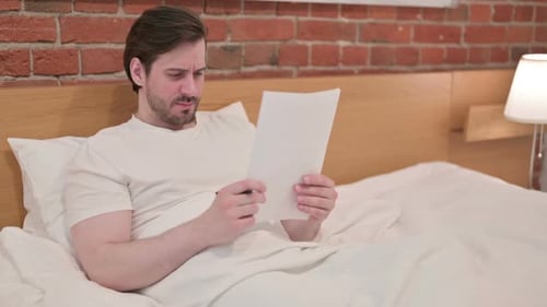 Man in Bed Reads Papers with Confused Expression