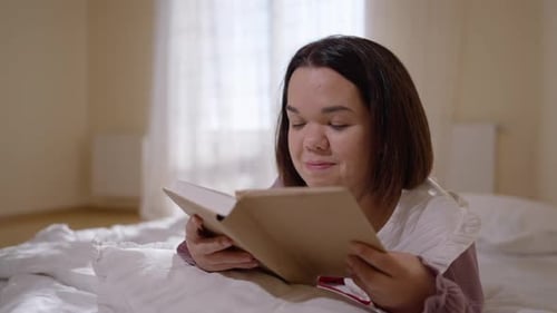 Young Happy Little Woman Reading Book Lying in Cozy Bed Smiling Looking Away