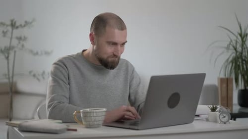Handsome young Caucasian man with beard in a gray jacket working at a laptop at home.