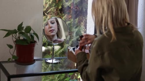 Beautiful blond hair woman in green shirt applying makeup in opposite of mirror