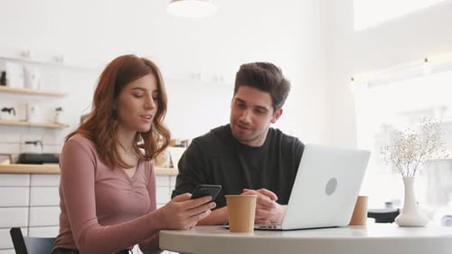 Two Young People Working with a Laptop and Smartphone in a Cafe and Drinking Coffee