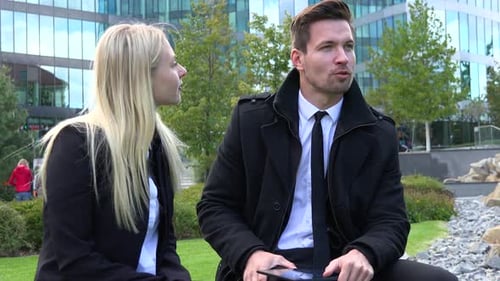 A Businessman and a Businesswoman Sit on a Bench and Talk in Front of an Office Building