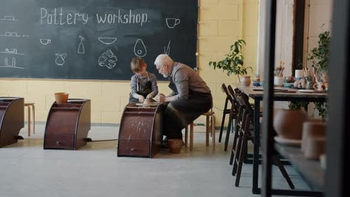Boy Working on Throwing Wheel While Senior Man Speaking Teaching in Pottery Workshop
