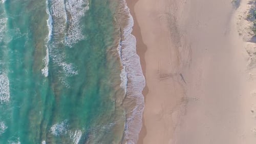 Aerial shot of waves crashing into sandy beach while person stands on shoreline