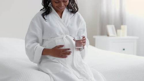 Positive, nicht wiederzuerkennende werdende afroamerikanische Frau, die ihren Bauch streichelt und Wasser trinkt von