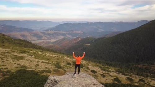 Aerial view of a hiker man raising his hands on a big rock in mountains.