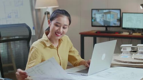 Asian Woman Engineer Typing On A Laptop And Looking At Blueprint Before Walking Out Of The Office
