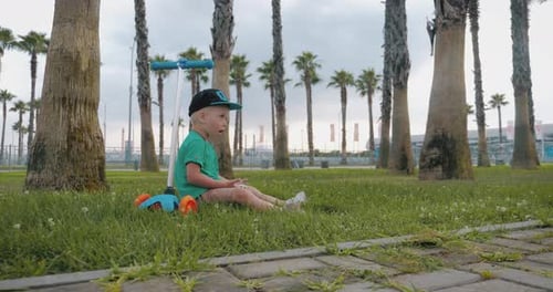 Young Boy Watches a Video on His Mobile Phone Sitting on the Grass in Park. Children and Modern