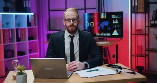 Businessman in Formal Wear in Glasses Sitting in front of Camera in Evening Office
