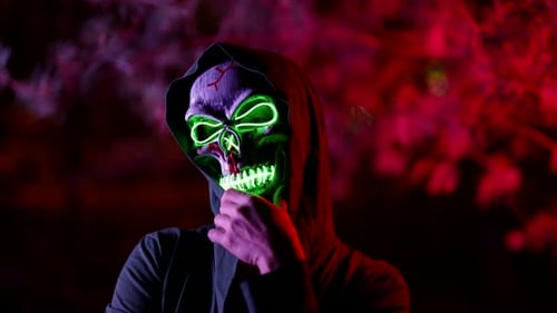 Enigmatic Figure of Person with Skull Mask on Face and Hood on Head Glowing in Darkness