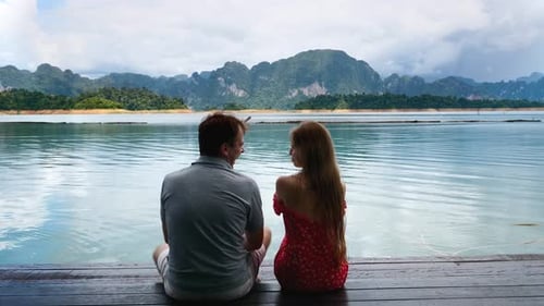 Romantic Pair of Man and Woman Sitting on Wooden Lake Pier with Green Mountains