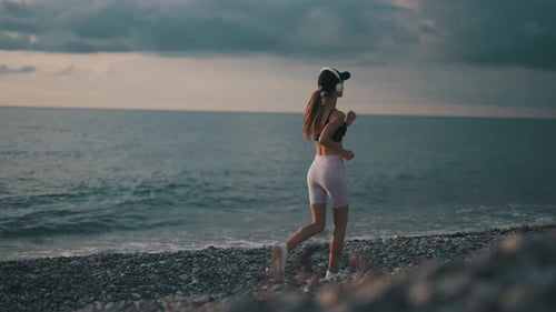A Young Woman Runner is Listening to Music in Earphones and Training By a Sea