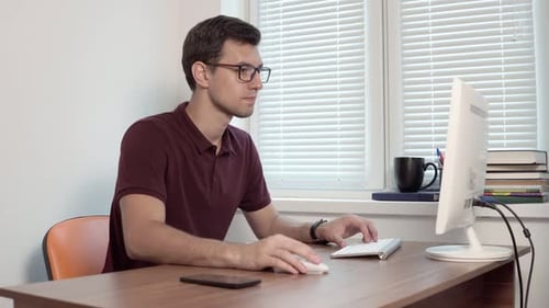 Junger Büroangestellter mit Brille sitzt im Büro am Fenster und tippt Bericht auf dem Computer