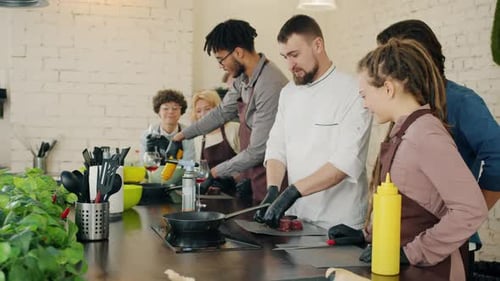 Männlicher Koch unterrichtet Gruppe von Auszubildenden beim Kochen von Fleisch und diskutiert Gerichte in der Küche