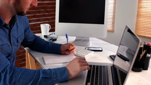 Man Works on Laptop and Writes in Notebook