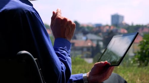 Old Senior Man Sits on the Bench in the Park and Works on Tablet - City in Background - Closeup