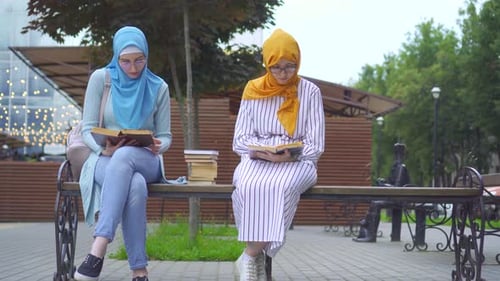 Zwei muslimische Studenten in traditionellen Schals mit Büchern in der Hand lesen auf einer Bank sitzend