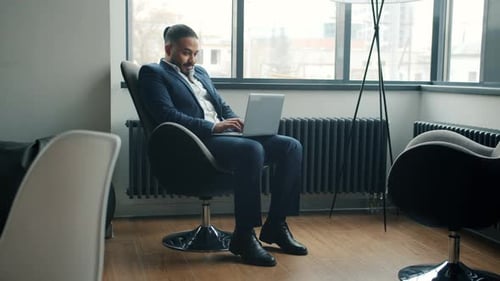 Cheerful Middle Eastern Businessman Using Laptop Typing Sitting in Armchair in Office Room