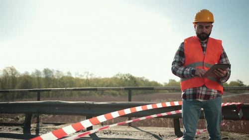 Young Bearded Man Inspector in an Orange Vest and Protective Helmet with Tablet