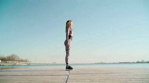 Young Fit Brunette Beautiful Girl in Leggings Black Sneakers Does Sport Exercises Against Blue Lake