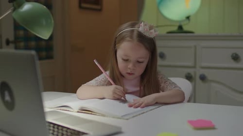 Close Up of Cute Preschool Girl Writing Down in Notebook with Pencil Looking at Laptop
