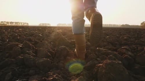 Farmer in Rubber Boots Walks Through a Plowed Agricultural Field