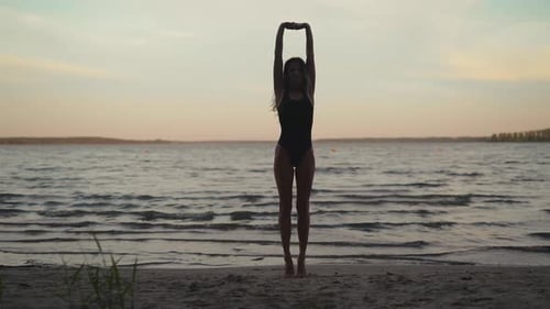 Silhouette Fit Woman Practices Yoga on a Sandy Beach Against Evening Lake