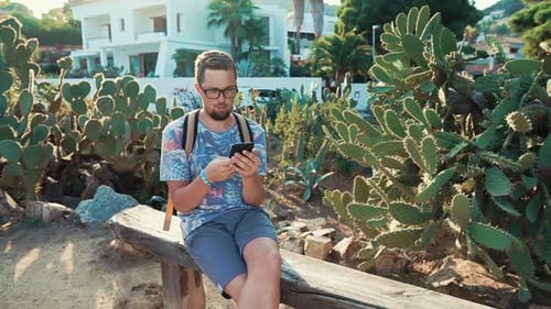 Man Is Sitting on a Bench in Garden Park in Sunny Evening, Using Smartphone